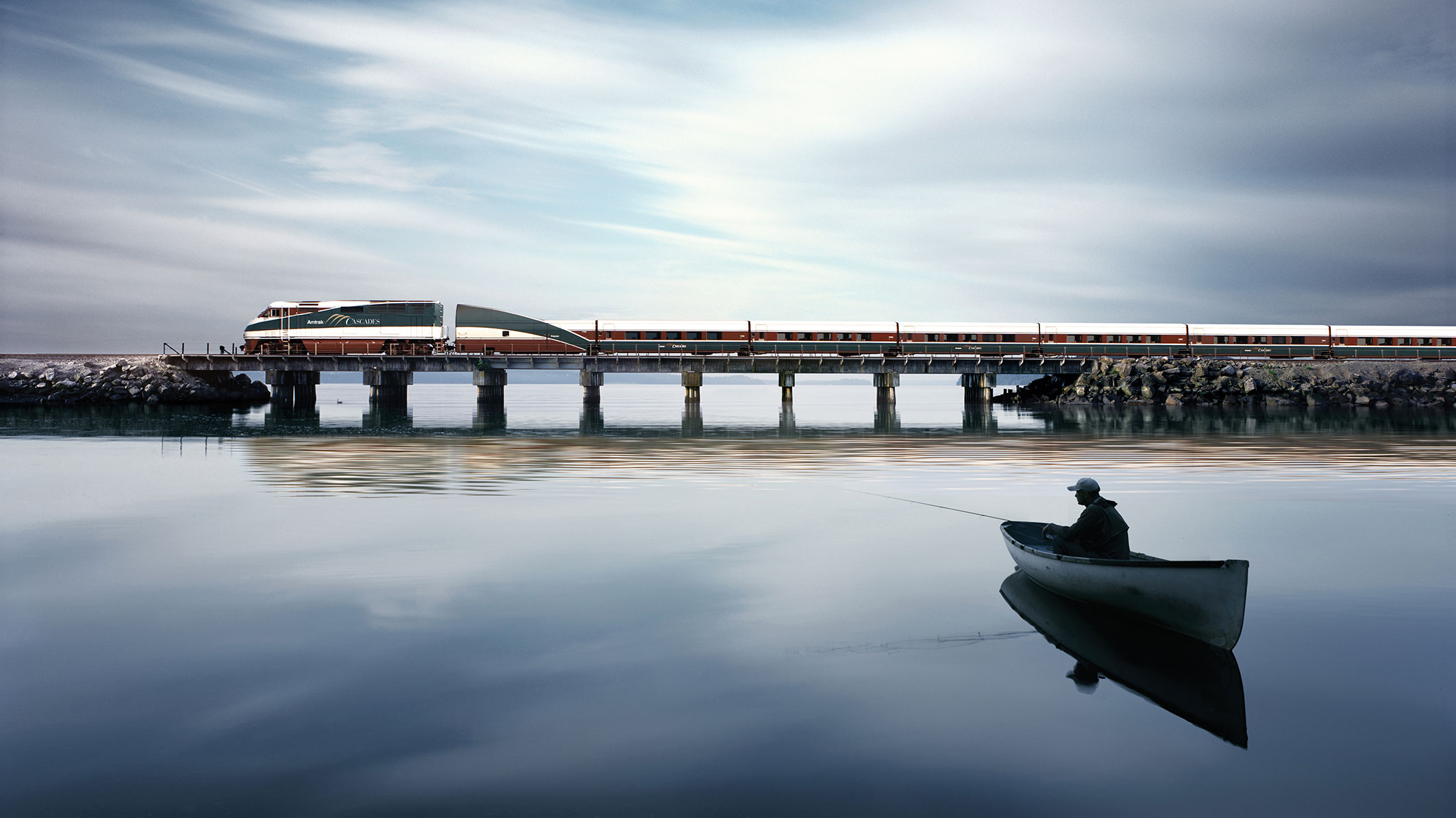 Amtrak Cascades Train Route train over water on a bridge with a man in a boat looking on