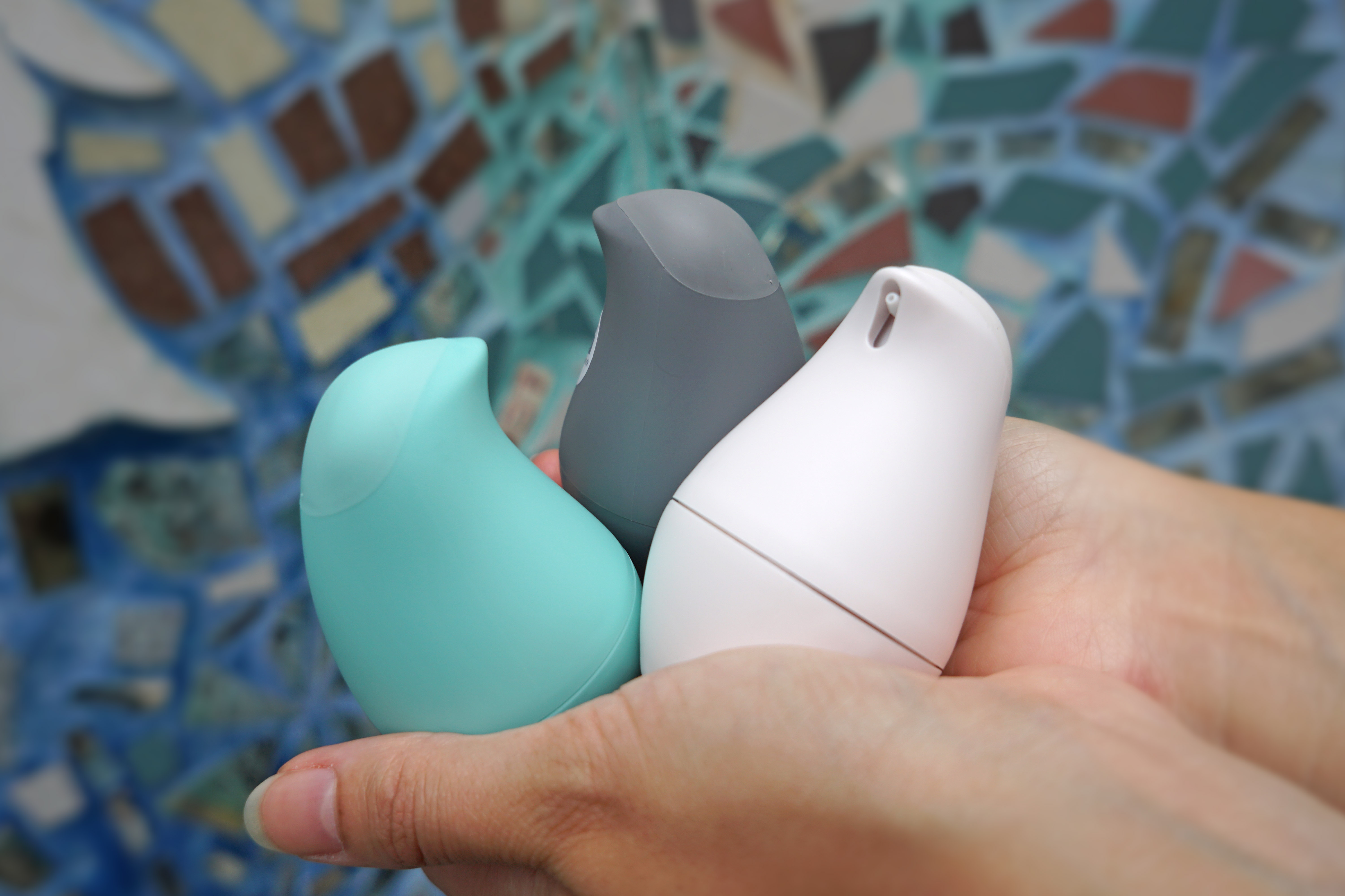 Holding all three colors of Olika Birdie hand sanitizers: blue, grey and white with mosaic background